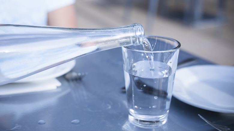 Slender clear bottle of water being poured into glass on metal table.