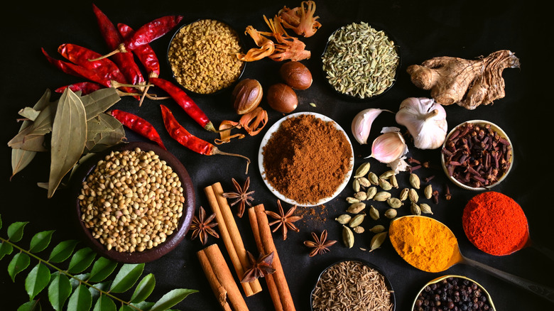 A spread of spices in a bowl, surround by traditional ingredients used for seasoning, like cinnamon sticks, chili, ginger, and garlic